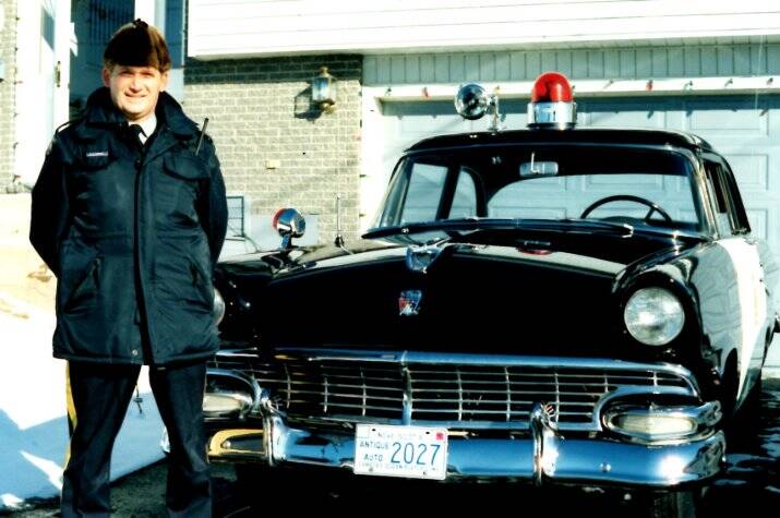 A dated picture of a uniformed RCMP officer standing next to a vintage police patrol car.