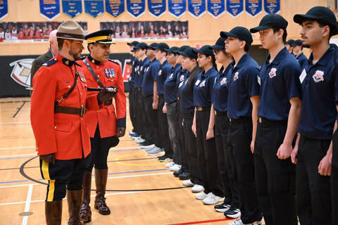Deux agents de la GRC vêtus de la tunique rouge se tiennent à côté d’une rangée d’élèves de la Youth Academy portant une casquette de baseball, une chemise à col bleu ornée d’un logo, un pantalon noir et des baskets dans un gymnase scolaire.